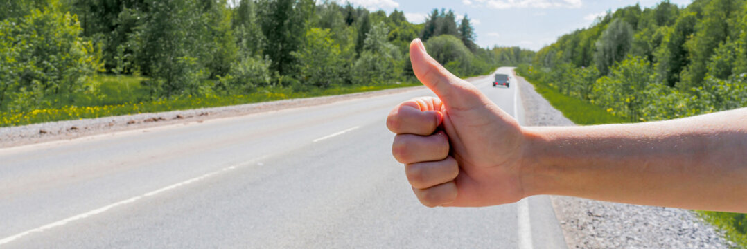 A man's hand and a hitchhiking sign on the road, hitchhiking around the city. The man tries to stop the car with his thumb up. Adventure and tourism. High quality photo