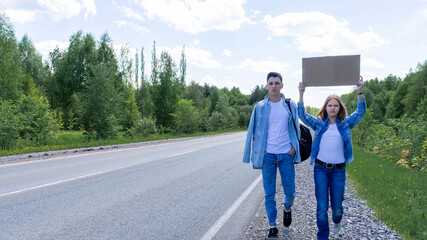 A young couple walks by the side of the road with a cardboard box in their hands with a place for text, hitchhiking, traveling, adventures. High quality photo