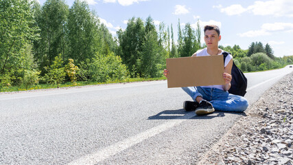 A young man is sitting on the side of the road with a cardboard sign in his hands. Space for text. Passion for travel, the concept of auto travel. A teenager tries to stop a car on the road with an