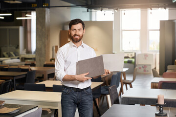 Front view of fashionable young male designing furniture indoors. Handsome brunette designer, seller standing, holding wooden samples, looking at camera. Concept of designing.