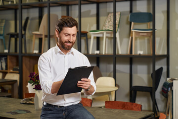 Front view of handsome brunette male with beard designing in furniture store. Designer, architect sitting on table, holding folder, writing, smiling. Concept of designing.
