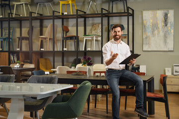 Front view of brunette male with beard designing in furniture store. Designer, architect sitting on...