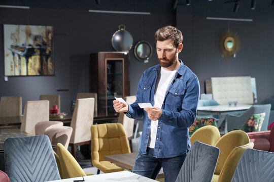Front view of brunette male choosing, buying furniture in store. Handsome boy with beard holding two cards, prices comprising, looking, thinking. Concept of urban lifestyle.