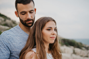 A close portrait of a Hispanic man who is hugging a Latina girlfriend on the rocky sea coast in Spain. A couple of tourists in the evening in a highland park in Valencia.