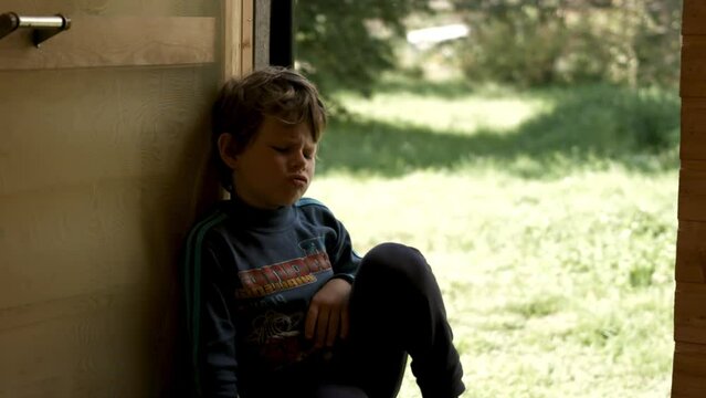 Boy child looking sad sitting by the opened door of a house with summer meadow behind. Creative. Little capricious boy make faces.