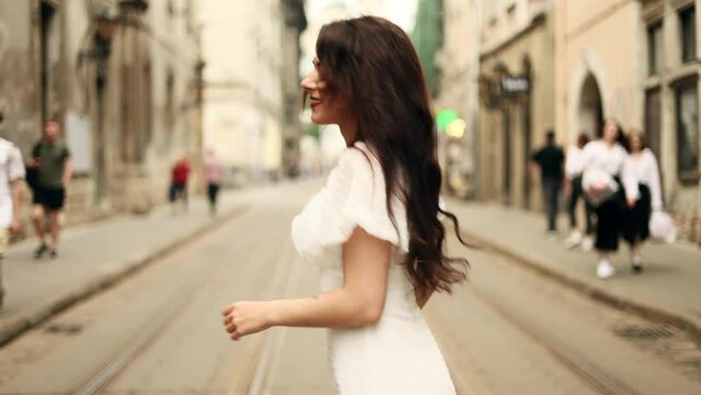 Smiling woman in white dress walking down the city street. Flirty girl with long dark hair turning around in city centre.