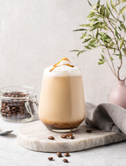 Cappuccino or latte with milk foam and caramel in a glass with coffee beans on light  marble background with branches. Front view and copy space