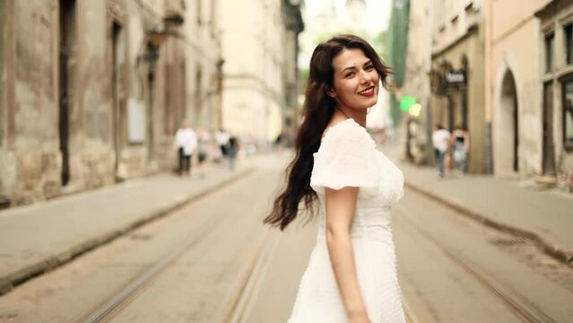 Smiling woman in white dress walking down the city street. Flirty girl with long dark hair turning around in city centre.