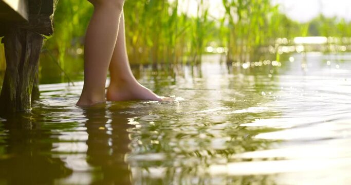Closeup of children legs which are swinging, while touching water in a lake in a relaxed way. A child is sitting on the edge of a wooden pier  in summer