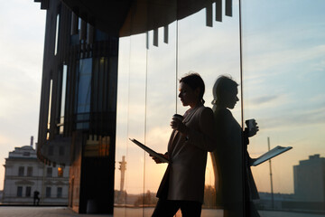 Busy contemporary lady in jacket standing by glassy building and drinking coffee while viewing papers outdoors, reflection effect