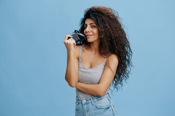 ONLINE GAME AD CONCEPT. Smiling beautiful curly Latin lady hold game pad near face, ready to play cool RPG, look at camera. Gaming accessories brands ad. Studio shoot isolated blue background