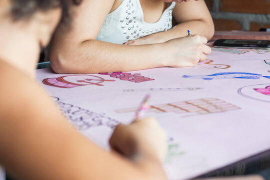 young latina woman helping her daughter to do her school homework, girl painting with her colored pencils over several drawings made on a pink cardboard. student concept.