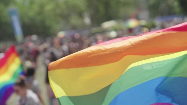 The LGBTQ rainbow flag waving in slow motion with people in a pride parade in the background