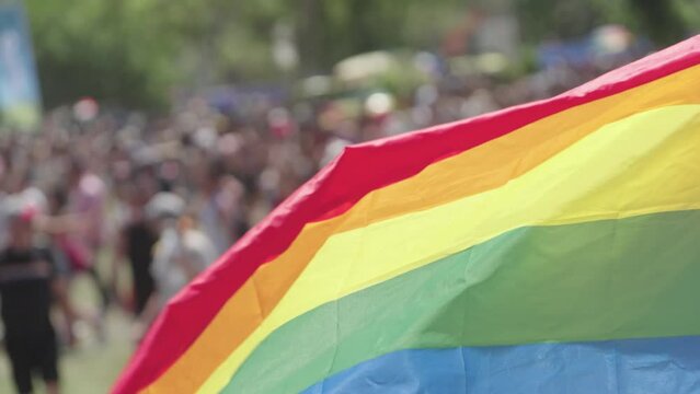 The LGBTQ rainbow flag waving in slow motion with people in a pride parade in the background