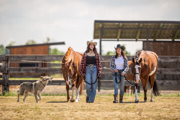 Two young female friends are walking their horses for a ride with their dog running around