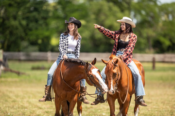 Two good-looking young women ride their horses, one pointing to the right while horses touch with...