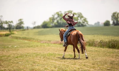 Schilderijen op glas Woman in jeans shirt holds her cowgirl hat as she gallops on a meadow on her paint horse © weyo