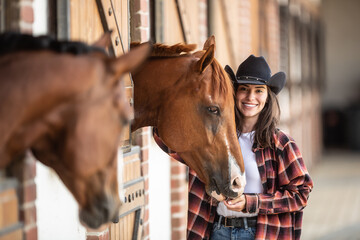 Good-looking girl in cowgirl hat stands next to horses inside the stable, smiling