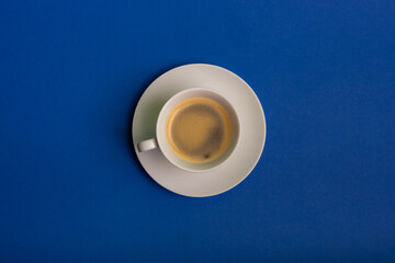 White cup of coffee with foam on round plate on blue background from above. Morning americano flat lay. Tasty hot coffee.