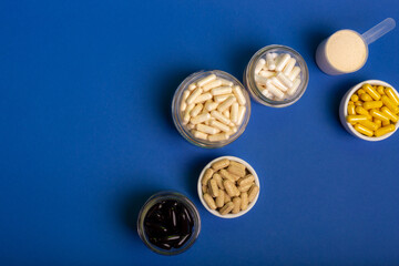 Black vitamin capsules, white vitamin capsules in glass jars and multivitamin tablets in small white plate from above on blue background.