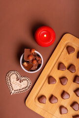 Heart shaped chocolate candies on wooden desk, milk chocolate with nuts on small round plate, wooden heart and red candle from above on brown background. Romantic holiday card with candies.