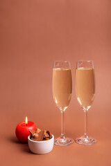 Two glasses of white sparkling wine, red candle and tasty milk chocolate with nuts in a small white plate on brown background. Beautiful holiday card.