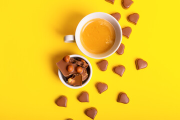 Heart shaped chocolate candies, milk chocolate with nuts on small round plate and a cup of coffee from above on yellow background. Romantic holiday card with candies. Valentine's Day breakfast.
