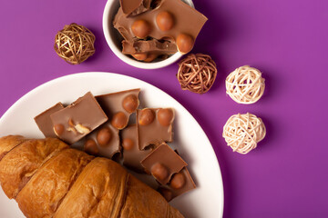 Sweet breakfast with morning hot coffee, tasty french croissant and sweet milk chocolate with nuts on white round plate from above on violet background. 