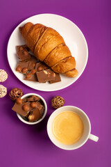 Sweet breakfast with morning hot coffee, tasty french croissant and sweet milk chocolate with nuts on white round plate from above on violet background. 