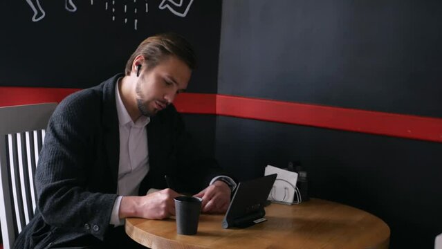 Handsome young smiling freelancer sitting at a caf&eacute; drinking coffee and talking on the tablet discussing and writing in notebook. Stylish buisnessman with headphones guarantees good deal for client
