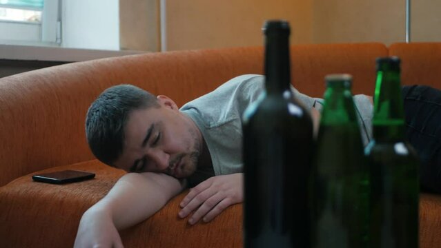 Young alcoholic waking up in the bed after alcohol abuse and drug addicted looking on the bottles with alcohol and starting to drink more. Alcoholism concept. Man drinking alcohol in the morning
