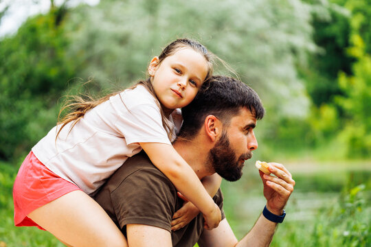 Side view of smiling family sitting on picnic in park forest around trees bushes. Little daughter with puffed cheeks sitting on fathers back. Middle-aged man eating food. Love, summer, togetherness.