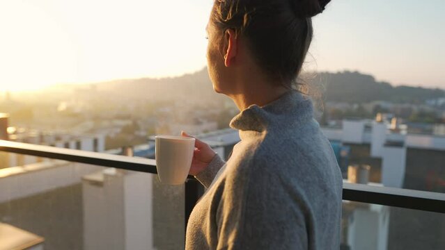 Woman starts her day with a cup of tea or coffee on the balcony at dawn, slow motion. City landscape in the background