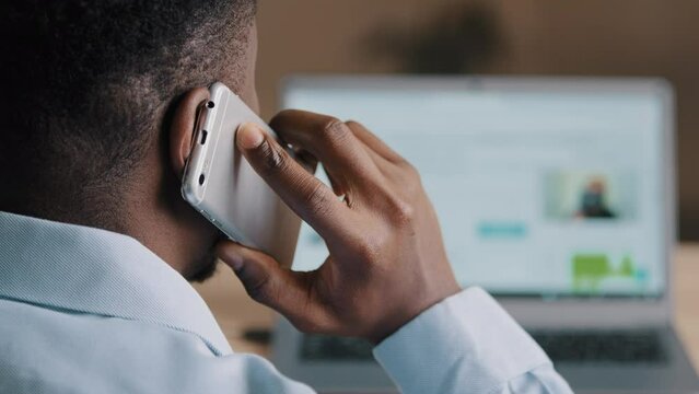 Backside unknown african american man young businessman web designer look at laptop screen surf internet website scroll page interface in social network talk mobile phone wireless call communication