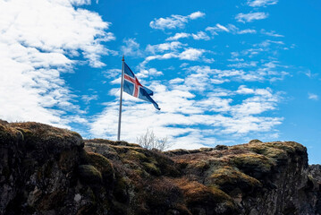 Beautiful Icelandic flag waving on rocky cliff. Low angle view of national flag on mountain against blue cloudy sky. Concept of patriotism in Thingvellir National Park.