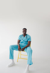 An African doctor in a turquoise medical suit sits on a chair on an isolated white background.