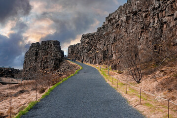 Fototapeta na wymiar Tourists walking on road amidst rock formations during sunset. Majestic cliffs in mountain against cloudy sky. People exploring dramatic landscape in Thingvellir national park.
