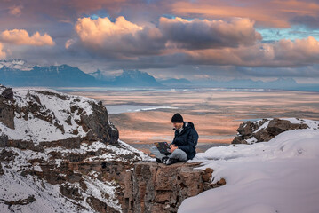 Young explorer using laptop while relaxing on snow covered cliff. Scenic view of volcanic landscape against cloudy sky. Male tourist using wireless technology during sunset. - Powered by Adobe