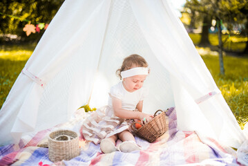 Little girl lying and playing in a tent, children's house wigwam in park Autumn portrait of cute curly girl. Harvest or Thanksgiving. autumn decor, party, picnic. Child in white dress with pumpkins.  © MartaKlos