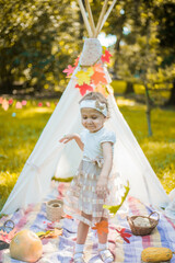 Little girl lying and playing in a tent, children's house wigwam in park Autumn portrait of cute curly girl. Harvest or Thanksgiving. autumn decor, party, picnic. Child in white dress with pumpkins.  © MartaKlos