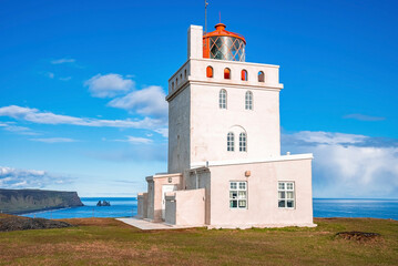 Fototapeta na wymiar Beautiful Dyrholaey lighthouse on cliff during sunny day. Idyllic view of white concrete building at seashore in Dyrholaeyjarviti. Scenic view of Reynisfjara beach against blue sky.