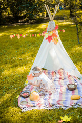 Little girl lying and playing in a tent, children's house wigwam in park Autumn portrait of cute curly girl. Harvest or Thanksgiving. autumn decor, party, picnic. Child in white dress with pumpkins.  © MartaKlos