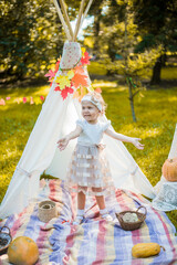 Little girl lying and playing in a tent, children's house wigwam in park Autumn portrait of cute curly girl. Harvest or Thanksgiving. autumn decor, party, picnic. Child in white dress with pumpkins.  © MartaKlos