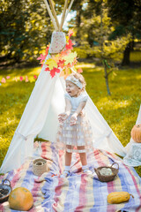 Little girl lying and playing in a tent, children's house wigwam in park Autumn portrait of cute curly girl. Harvest or Thanksgiving. autumn decor, party, picnic. Child in white dress with pumpkins.  © MartaKlos