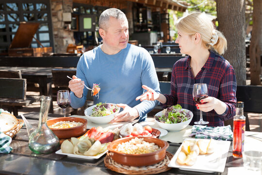 Family Couple Enjoying Warm Spring Day And Delicious Dinner With Wine In Cosy Country Restaurant In Open Air