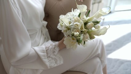 The bride in pajamas holds a bouquet. Morning of the bride, the bride in white pajamas holds a light wedding bouquet.