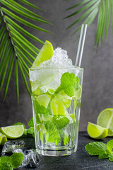 Non-alcoholic mojito with crushed ice and lime on top of glass with transparent straw. Fresh drink stands on table near slices of fruit and mint leaves closeup
