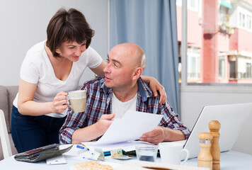 Portrait of man working with financial documents at laptop, woman offering coffee to him