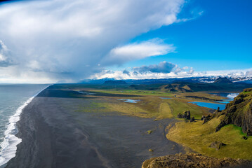 Idyllic view of black sand beach by mountains. High angle view of Dyrholaeyjarviti cliff. Beautiful picturesque scenery of volcanic landscape against blue cloudy sky .