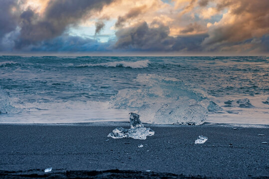 Close-up of iceberg on shore at Diamond beach. Chunk of floating ice from Jokulsarlon glacier lagoon on black sand. Beautiful view of waves rushing at shore against cloudy sky during sunset.